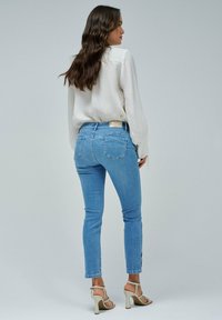 Jeans de mezclilla azul claro con un corte ajustado, que cuentan con bolsillos traseros y un ligero estiramiento; combinados con una blusa blanca texturizada y tacones beige.