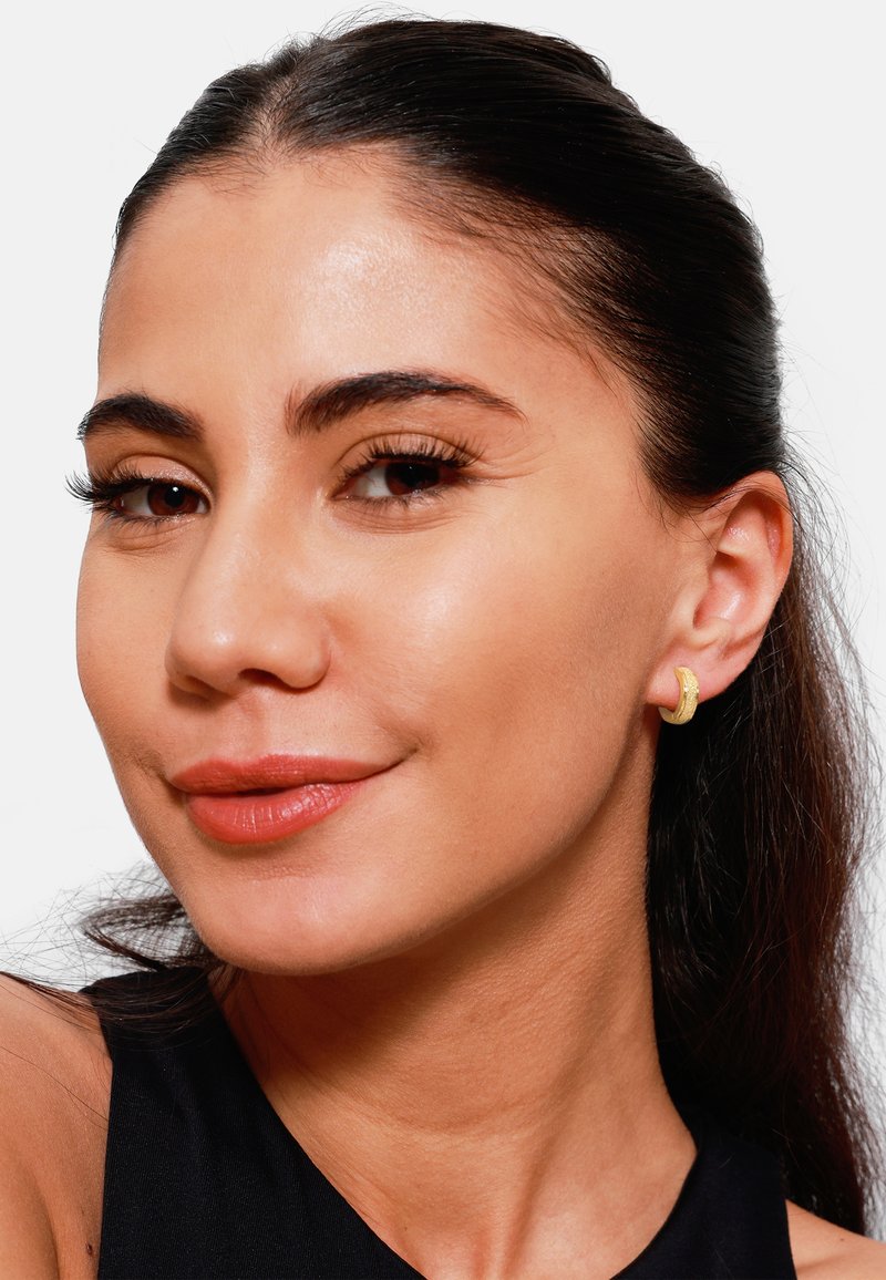 Boucles d'oreilles en or avec une finition texturée, portées à l'oreille droite. Le modèle a les cheveux foncés et porte une couleur de lèvres neutre, sur un fond blanc.