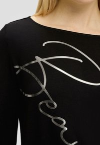 Czarna koszula z długim rękawem i szerokim dekoltem, zdobiona metallicznymi, srebrnymi abstrakcyjnymi wzorami na przodzie. Miękka, gładka tekstura materiału.