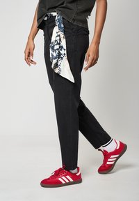 Pantalones negros de pierna recta con una textura suave, combinados con zapatillas rojas con rayas blancas y una bufanda estampada atada a la cintura.