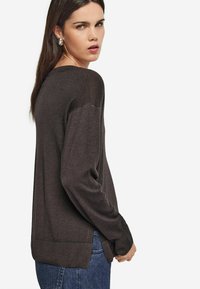 Kvinna med långt mörkt hår bär en mörkbrun tröja och blåjeans, står vänd åt sidan med huvudet vänt mot kameran mot en vit bakgrund.