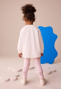 White long-sleeve shirt, pink leggings with cartoon sheep and clouds, white shoes, standing with a blue abstract shape in background.