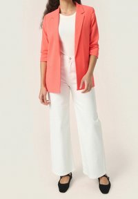 Koralowy blezer o dopasowanym kroju z podwiniętymi rękawami, zestawiony z białymi, szerokimi spodniami i czarnymi balerinami. Gładkie tekstury w całym stroju.