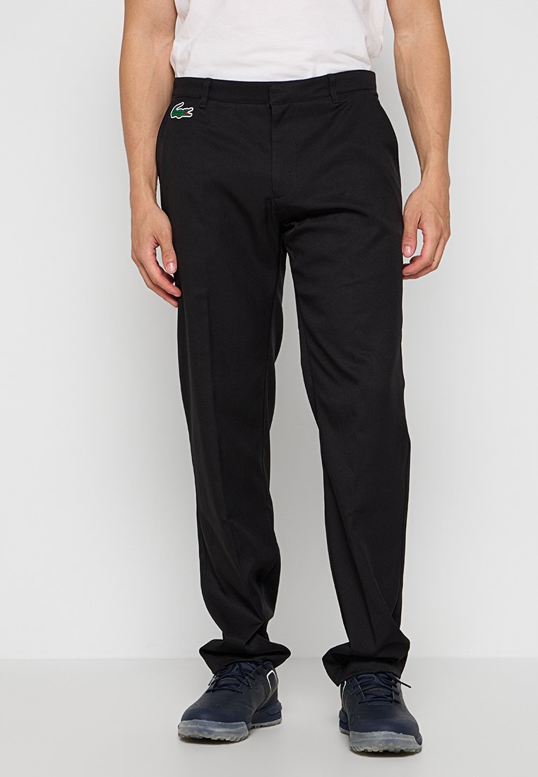 Pantalon noir en tissu lisse, design ajusté, coupe droite, avec un petit logo vert Lacoste sur la zone de la poche gauche.
