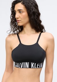 Γυναίκα με σγουρά μαλλιά που κοιτάζει αριστερά, φοράει αθλητικό σουτιέν Calvin Klein σε μαύρο χρώμα με λεπτούς τιράντες και ελαστική ταινία με το λογότυπο.