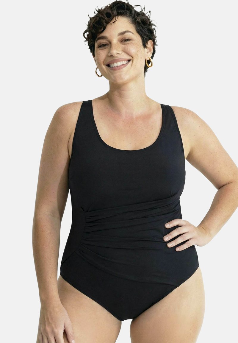 Femme souriante aux cheveux courts et bouclés portant un maillot de bain une pièce noir froncé, debout avec une main sur la hanche devant un fond uni.