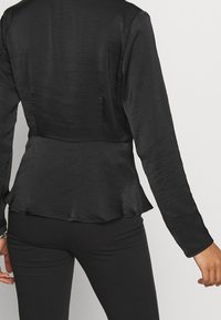 Černý peplum top z lehké, texturované tkaniny. Dlouhé rukávy, přiléhavý korzet a volánkový detail na spodním lemu. Spárováno s černými kalhotami.