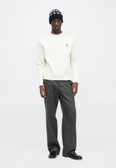Man wearing a white ribbed sweater with a fox patch, wide gray trousers, black leather shoes, and a navy knit beanie with white patterns.