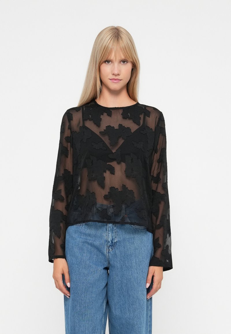 Sort gennemsigtig bluse med lange ærmer og blomsterbroderi. Har en rund halsudskæring og et cropped design, som er parret med blå bredbenede jeans.