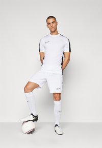 Completo sportivo Nike in bianco, composto da una maglietta a maniche corte con dettagli neri e pantaloni corti abbinati. Giocatore che indossa calze bianche e scarpini, in piedi su una palla.