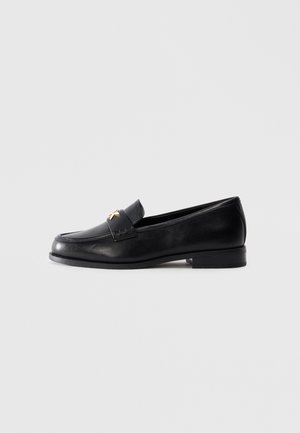 Mocassino in pelle nera con tacco basso e emblema decorativo dorato sulla fascia, esposto su uno sfondo grigio chiaro uniforme.