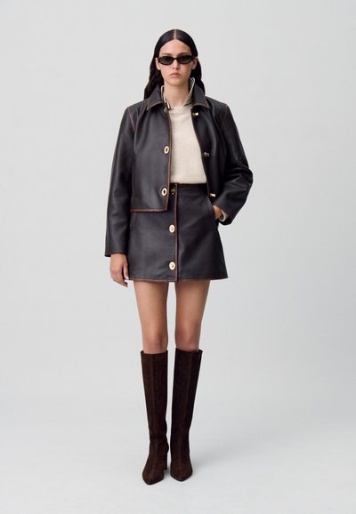Giacca di pelle marrone con cuciture a contrasto, bottoni dorati e una mini gonna abbinata. La modella indossa stivali in suede marrone fino al ginocchio.