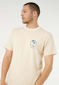 T-shirt beige in cotone con grafica blu di palme e scritta "RIPCURL" all'interno di un motivo circolare sul lato sinistro del petto. Maniche corte.