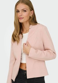 Femme aux longs cheveux blonds portant des boucles d'oreilles créoles dorées, un blazer rose clair, un crop top blanc et un jean noir, posant devant un fond uni.