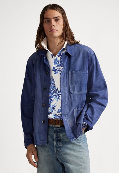 Μπλε βαμβακερό μπουφάν με κουμπιά μπροστά, μεγάλο τσεπάκι στο στήθος και χαλαρή γραμμή. Φοριέται πάνω από πουκάμισο με λευκό και μπλε σχέδιο.