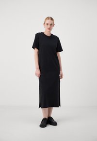 Black cotton dress with short sleeves, straight cut, side slits, and a crew neckline. Paired with black shoes. Plain texture.