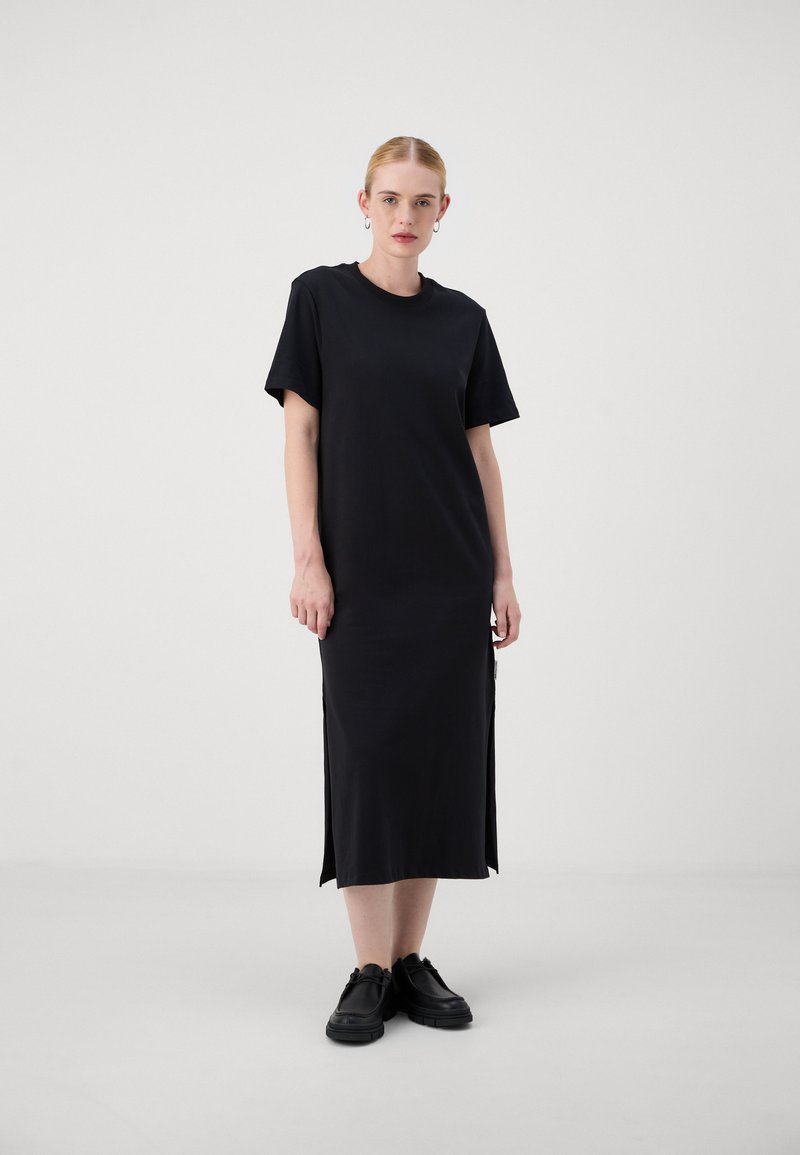 Black cotton dress with short sleeves, straight cut, side slits, and a crew neckline. Paired with black shoes. Plain texture.