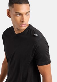 Camiseta negra de manga corta hecha de tela suave, con cuello redondo y una pequeña etiqueta de logo en el hombro, con detalles de costura visibles.