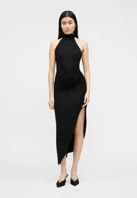 Femme en robe noire sans manches à col montant, avec un ourlet asymétrique et une fente latérale, portant des chaussures à talons pointues noires, debout devant un fond neutre.