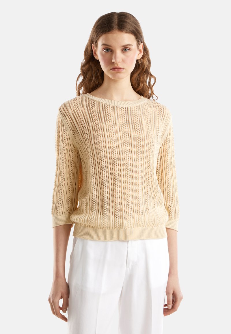 Jasno beżowy sweter wykonany na drutach, o luźnym kroju, z szerokim dekoltem, rękawami o długości trzech kwart i naprzemiennymi wzorami otwartymi i zamkniętymi.
