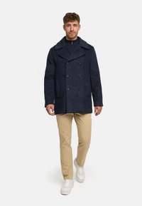 Abrigo de lana azul marino con diseño de doble botonadura, grandes solapas y dos bolsillos frontales, llevado sobre pantalones beige y zapatillas blancas.