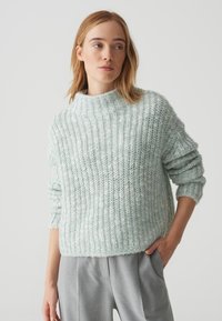 Maglione azzurro chiaro lavorato a maglia con colletto alto, texture a coste e spalle scese. Abbinato a pantaloni grigi, mani nelle tasche.