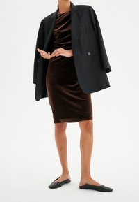 Brun sammetsklänning i kombination med en överdimensionerad svart blazer, ankellång och platta svarta skor; släta texturer och modern silhuett.