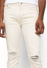 Pantalones vaqueros de mezclilla beige con desgarros en las rodillas, diseño de cinco bolsillos y cierre de botón. Textura suave y corte recto.