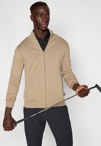 Μπεζ πουλόβερ με φερμουάρ σε υφή πλέξης, με μακριά μανίκια και γιακά όρθιο. Κρατώντας μπαστούνι γκολφ σε ένα χέρι με σκούρο παντελόνι.