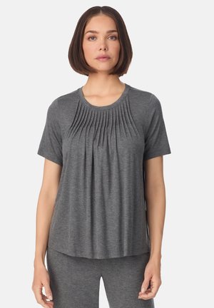 Mujer vistiendo una blusa gris plisada de manga corta y pantalones grises a juego, de pie contra un fondo claro y liso.