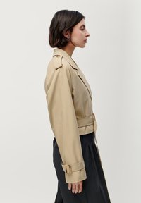 Beige trenchcoat met een tailleriem, brede revers en grote mouwdetails. De stof lijkt glad met een lichte glans. Gepresenteerd op een model.