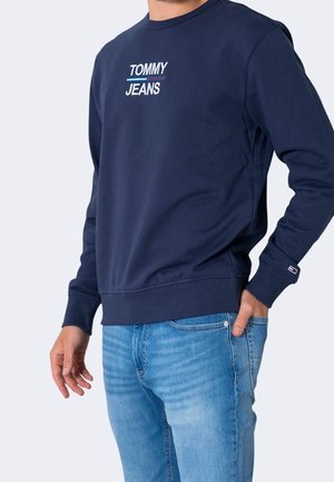 Man wearing navy blue Tommy Jeans sweatshirt and light blue jeans, standing with one hand in pocket against plain background.
