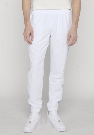 Mand iført hvide joggerbukser med elastisk talje, blå sidelister og hvide sneakers, stående mod en ensfarvet baggrund.