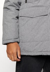 Graue gepolsterte Jacke mit einem strukturierten Stoff, umklappbaren Manschetten und einer Brusttasche. Hand ruht auf dem Kleidungsstück, sichtbare Nähdetails.