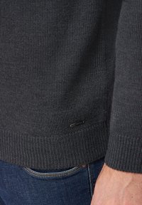 Grau gestrickter Pullover mit Rippstruktur und Bündchen. Mit einem kleinen Markenetikett am Saum, getragen über dunkelblauen Jeans.