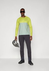 Mand iført sorte solbriller, limegrøn og lyseblå langærmet skjorte, sorte bukser, sorte sneakers, holder en grå cykelhjelm.