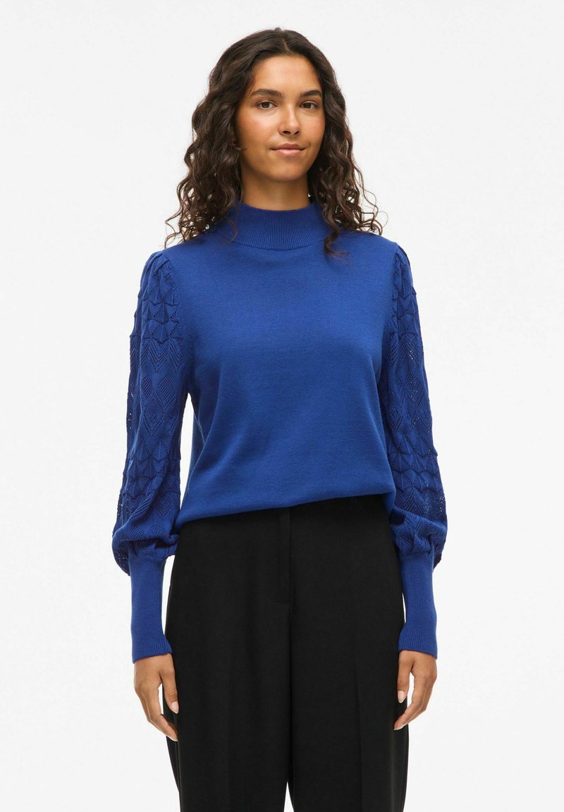 Königsblaues Pullover-Sweatshirt mit hohem Kragen, mit strukturierten gestrickten Puffärmeln und einem glatten Körper. Kombiniert mit schwarzen Hosen.