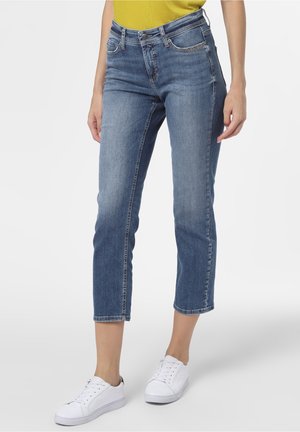 Femme portant un jean bleu taille mi-haute, coupe slim, raccourci au-dessus des chevilles, associé à des baskets blanches et un top jaune, debout sur un fond uni.