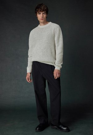 Strickpullover in hellbeige mit einem strukturierten Muster, kombiniert mit lockeren schwarzen Hosen und schwarzen Schuhen. Einfacher runder Ausschnitt, Ärmel mit Bündchen.