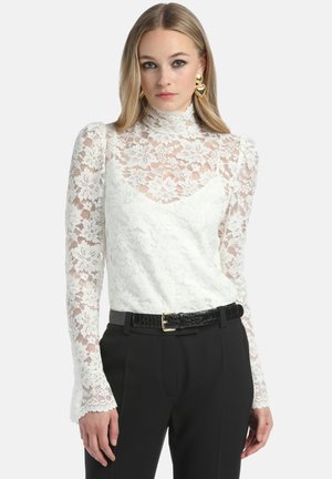 Witte kanten top met lange, doorschijnende mouwen met bloemenpatronen, een hoge hals en een aansluitende lijfje, gedragen met zwarte broek en een riem.
