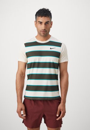Ein Mann trägt ein weißes und dunkelgrün gestreiftes Nike-T-Shirt mit blauen Akzenten und kastanienbraunen Shorts, steht vor einem schlichten weißen Hintergrund.