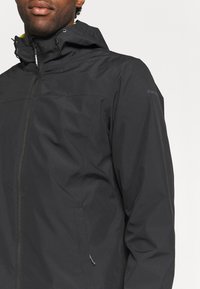 Homme portant une veste noire à capuche avec une fermeture éclair à l'avant et des cordons réglables sur la capuche, sur fond blanc uni.