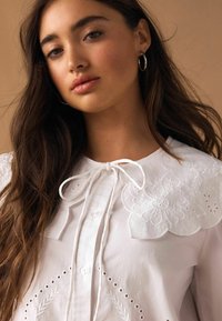 Fehér blúz virágos hímzett gallérral és megkötővel. Gombos záródással, lyukacsos részletekkel és sima pamut anyaggal rendelkezik.