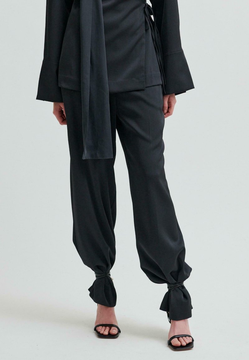 Czarne spodnie o dopasowanym kroju z szerokimi nogawkami, przylegające w okolicy kostek, zabezpieczone na sznurki. W zestawie z dopasowaną bluzką z długim rękawem i sandałami.