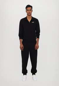 Sort zip-up sweatshirt med krave og manchetter, parret med løse sorte sweatpants og hvide sneakers. EA7-logo på brystet.