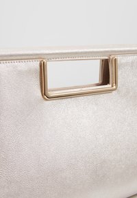 Metallinhohtoinen hopeanvärinen käsilaukku, jossa on teksturoidun pinnan lisäksi suorakulmainen kullanvärinen metallikahva ja selkeät linjat.