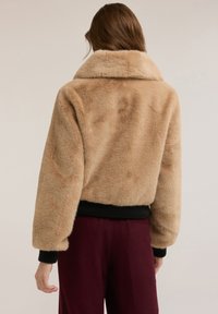 Kobieta ubrana w beżową kurtkę z futerkiem ekologicznego z czarnymi ściągaczami na rękawach i w pasie, w połączeniu z ciemnoczerwonymi spodniami, widziana od tyłu.