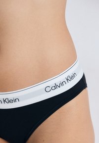 Nærbilde av en persons hofte iført svarte Calvin Klein underwear med en hvit midjebånd og svart logotekst.
