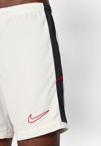 Bijele atletske šortsice s crnim bočnim panelima i crvenim Nike logotipom na donjoj lijevoj nozi koje nosi osoba.