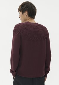 Ein Mann, der von hinten zu sehen ist, trägt einen burgunderfarbenen Strickpullover mit einem strukturierten Einsatzmuster und schwarze Hosen vor einem weißen Hintergrund.